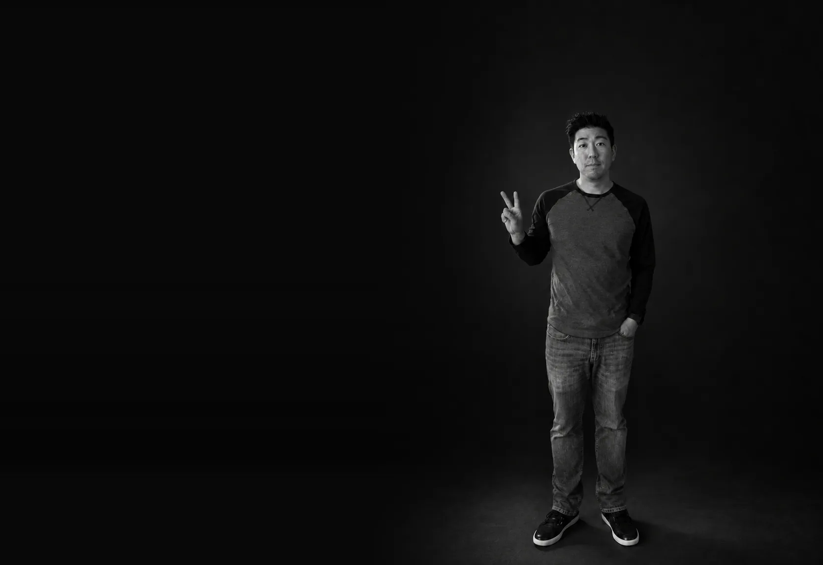 Black-and-white photo of Daniel Okamura, standing, flashing a peace sign